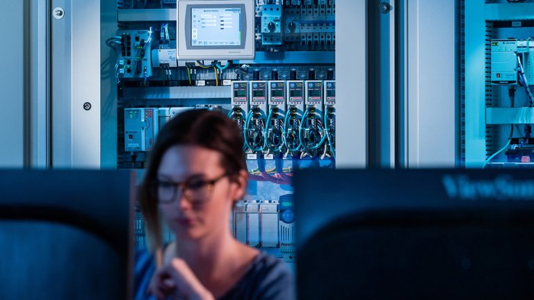 前景中有一位女性工程師正在電腦前工作，而背景中有裝滿 Kinetix 伺服驅動器的電氣機櫃與其他 Allen-Bradley 元件。