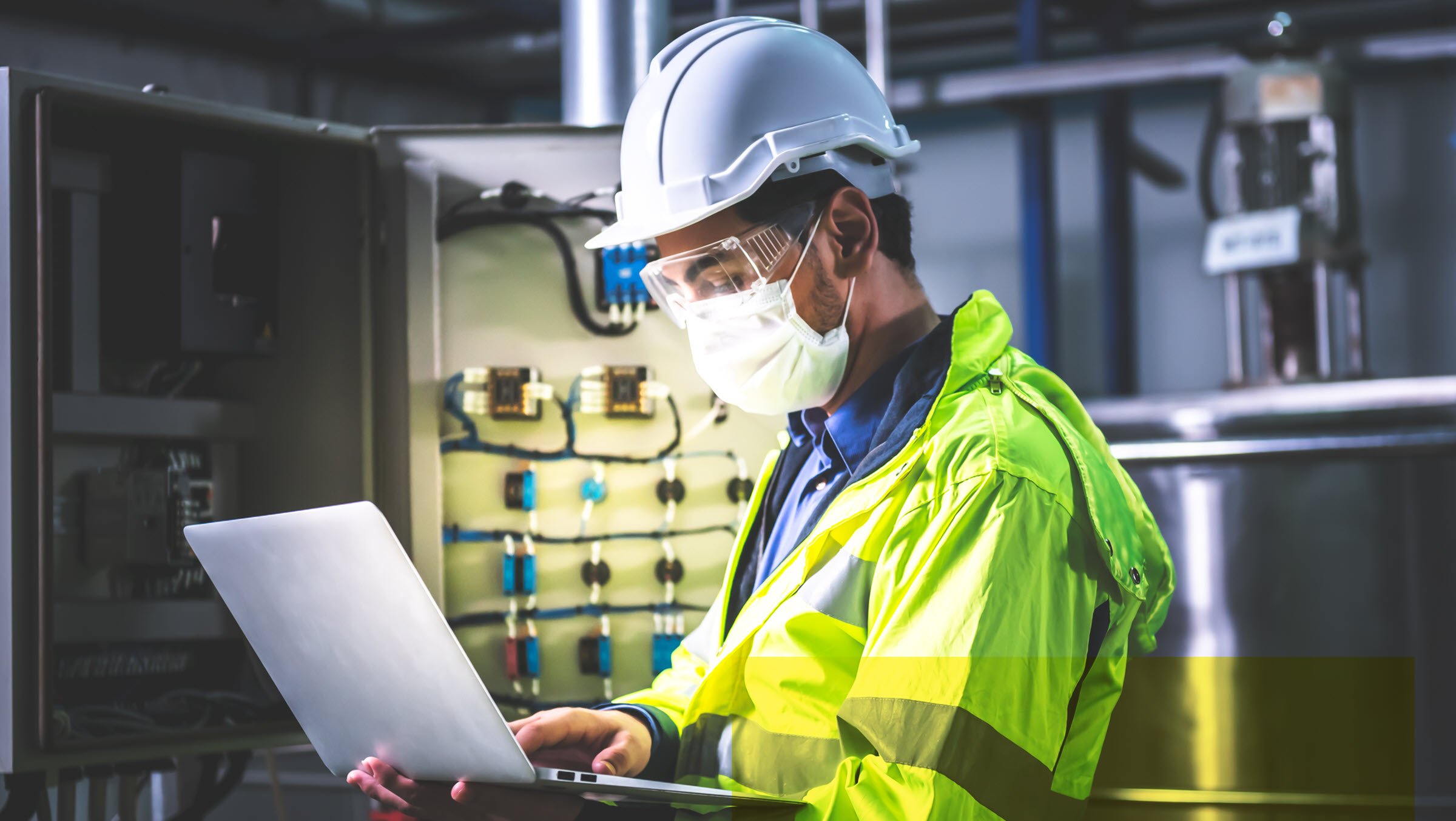 Portrait of a technician in hardhat and surgical mask is using computer notebook to check the electrical system of the machine in an industrial factory