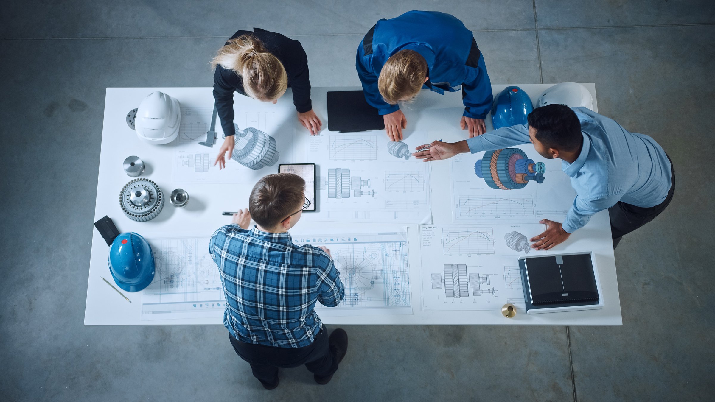 Équipe d’ingénieurs industriels penchés sur un bureau, analysant des plans de machines, résolvant des problèmes d’architecture, consultant un projet sur une tablette et inspectant un composant métallique. Vue de dessus à plat