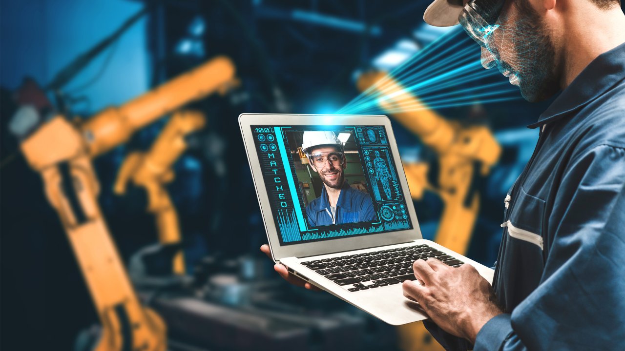 头戴安全帽的工人使用面部识别技术通过笔记本电脑远程控制工业机器。