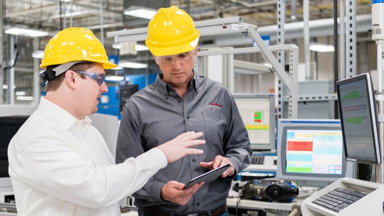 Dos trabajadores con cascos de seguridad amarillos en una fábrica moderna discuten datos en una tableta.
