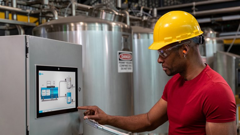 Eine Person interagiert mit einem HMI-Bedienfeld, das an einer Industrieanlage angebracht ist. In der einen Hand hält er ein Tablet