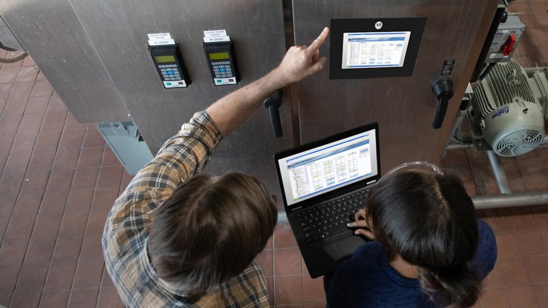 Une personne fait un geste vers un panneau sur les machines industrielles tandis qu'une deuxième personne observe, une console de programmation portable à la main.