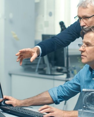 Ein Mitarbeiter sitzt an einem Schreibtisch und betrachtet zusammen mit einem anderen Mitarbeiter, der über die Schulter des anderen gebeugt auf Informationen auf dem Bildschirm deutet, einen Computer