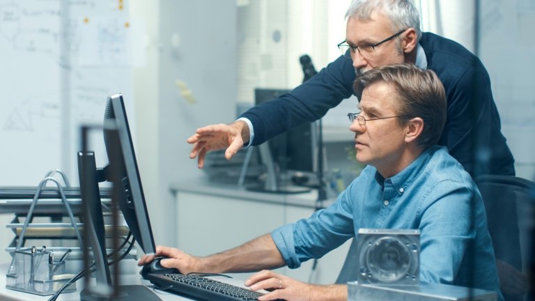 Um funcionário sentado em uma estação de trabalho e examinando um computador com outro funcionário de pé e curvado atrás dele, apontando para informações na tela.