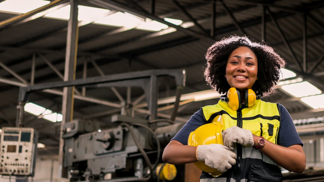 Engenheira em pé de frente às máquinas em uma fábrica, usando colete de segurança amarelo, fones de ouvido e capacete