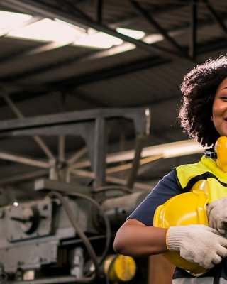Ingegnere donna in fabbrica, con indosso una tuta gialla, cuffie e casco, che sta in piedi di fronte a un macchinario