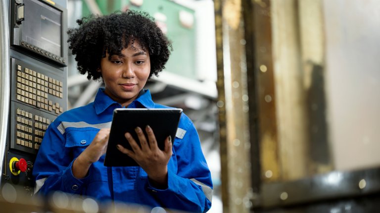 Processo di impostazione di una fresatrice CNC. Tecnico donna afroamericana prepara la macchina in base allo standard di lavorazione. Sta usando un tablet in una linea di produzione.