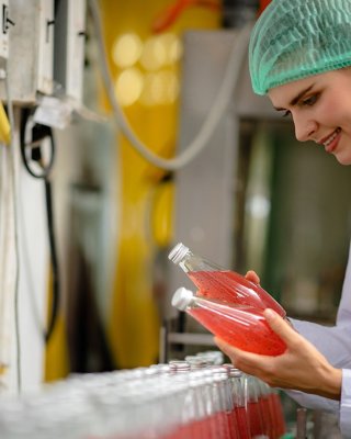 Woman wearing hair net and sterile clothing standing in front of conveyor line holding two glass bottles filled with pink liquid.