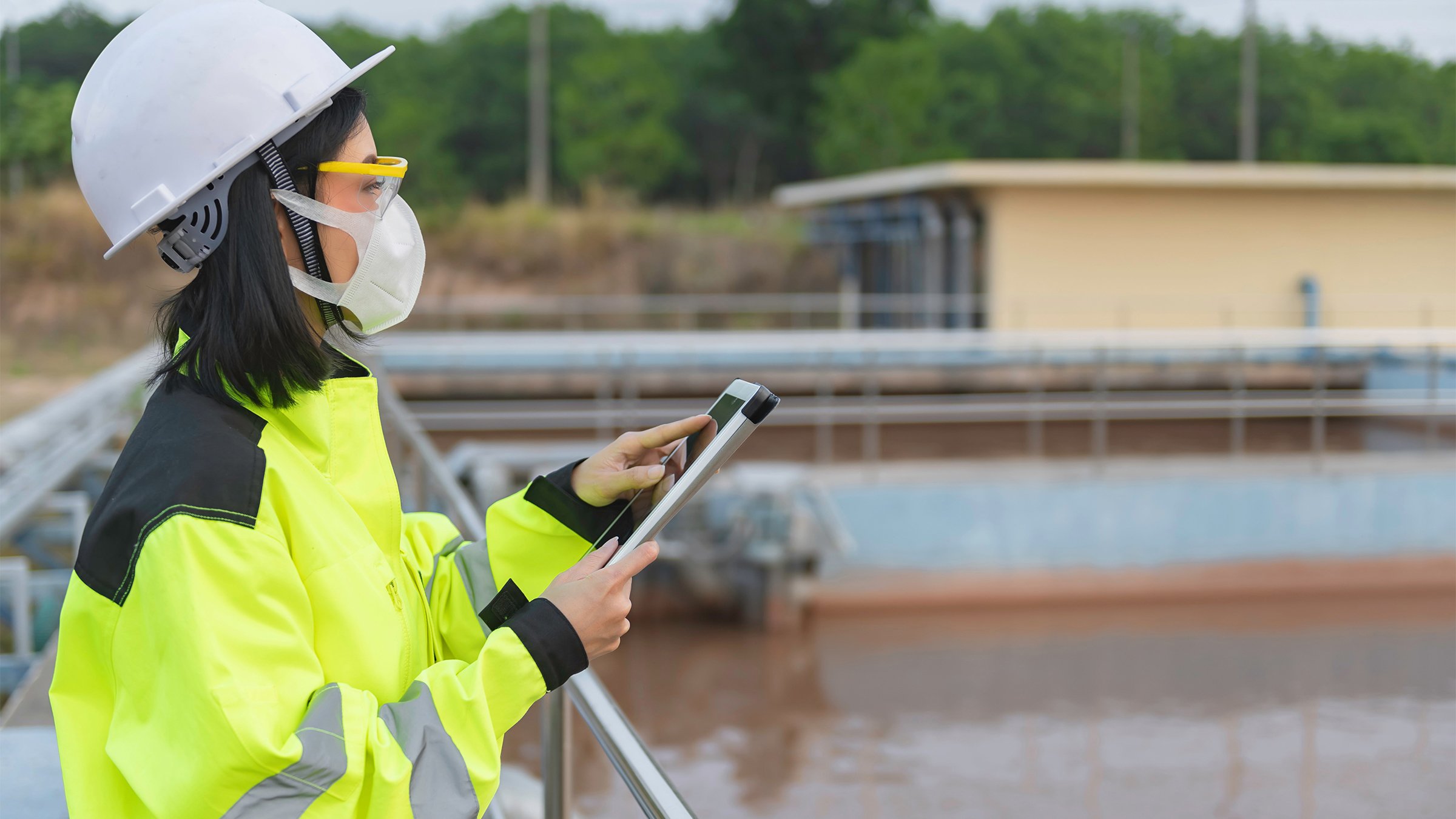Woman wearing protective gear using a tablet outdoors at a water treatment plant.