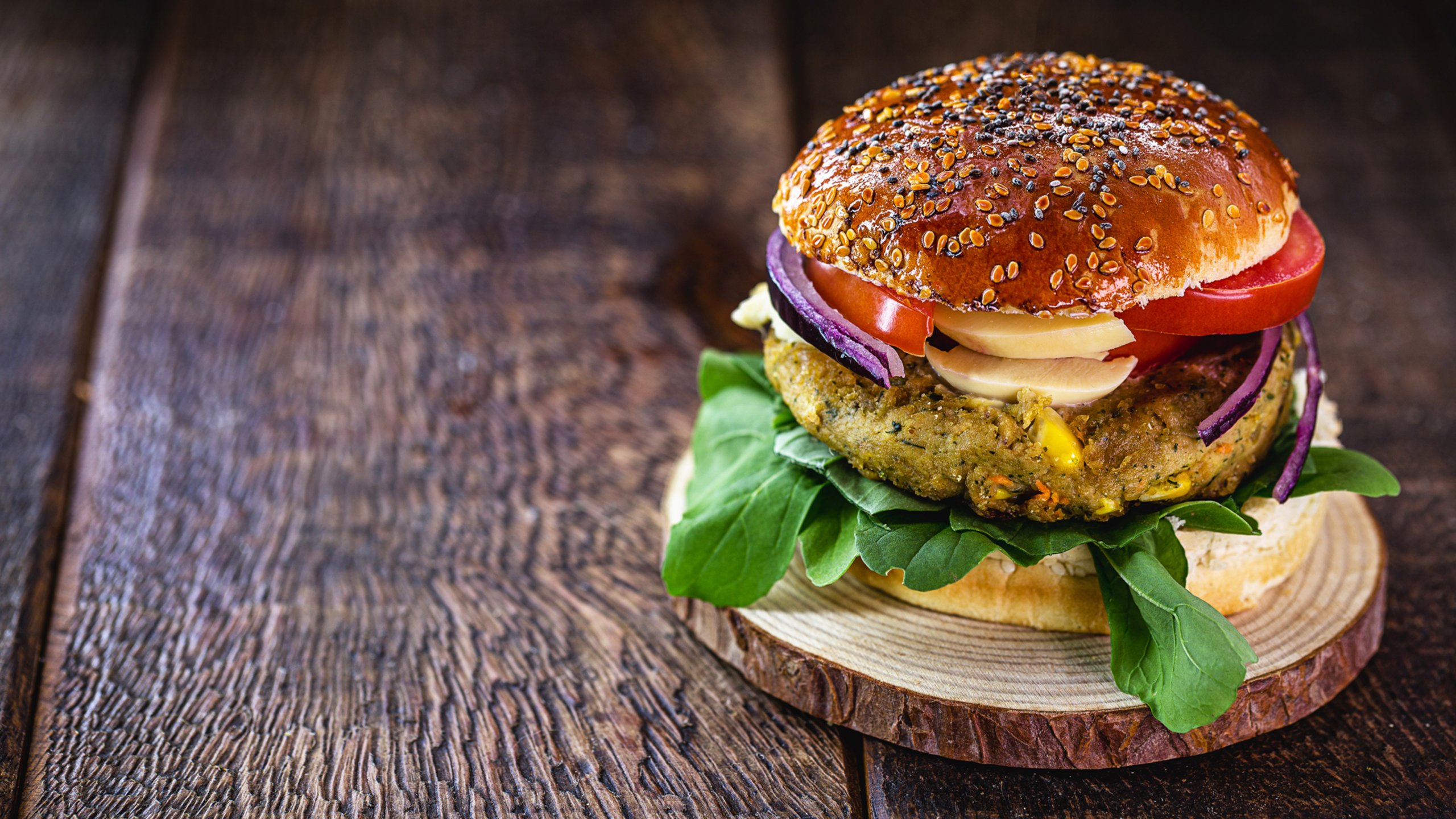 Plant-based meat burger on a sesame seed bun with tomatoes, mushrooms, onions and lettuce. The burger is on a wooden plate, sitting on a wooden table. The burger is on the right side of the image.