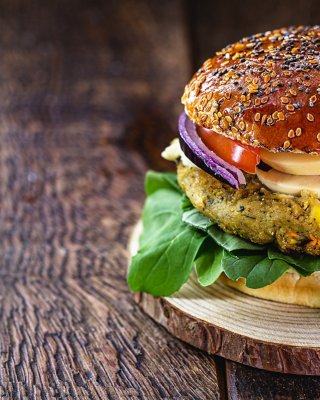 Burger mit pflanzenbasiertem Fleisch auf einem Sesambrötchen mit Tomaten, Pilzen, Zwiebeln und Salat. Der Burger liegt auf einer Holzplatte, die sich auf einem Holztisch befindet. Der Burger ist rechts im Bild.