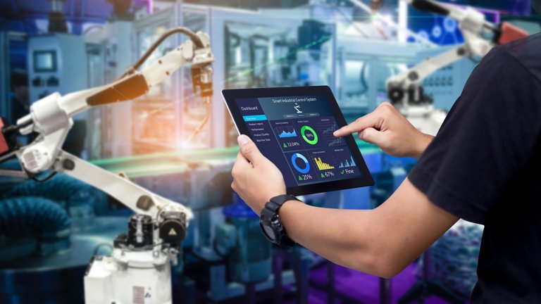 Vue en gros plan des mains d’un technicien tenant une tablette montrant des analyses dans une usine devant un bras robotisé blanc.