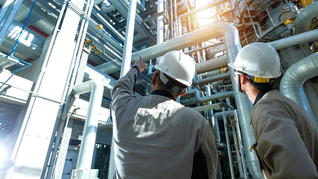 Dos trabajadores con cascos en una refinería de petróleo y gas forman una zona industrial. Detrás se ve el amanecer con un cielo nublado