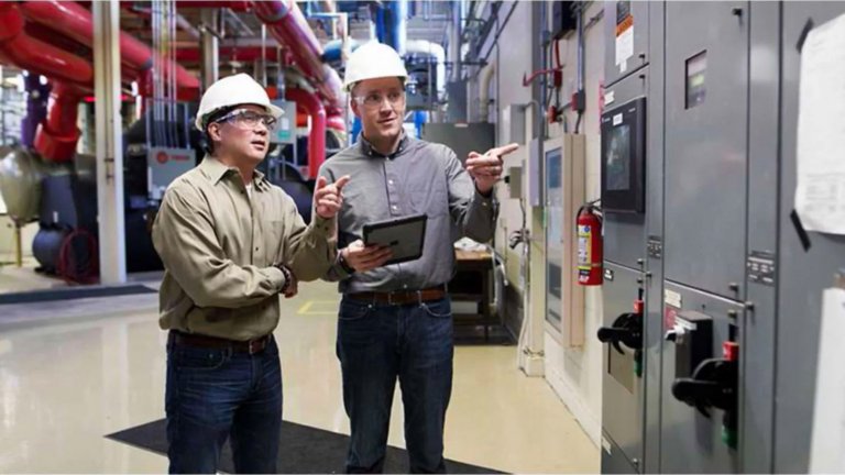Two men wearing hard hats looking at and pointing to a monitor. One man is holding a tablet They are in an Intelligent Packaged Power IPP factory.