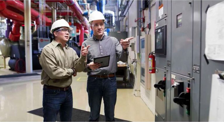 Two male engineers wearing hardhats inside a factory are looking at  a CENTERLINE motor control center, a grey metal cabinet with multiple doors, knobs and buttons that housed electrical components.
