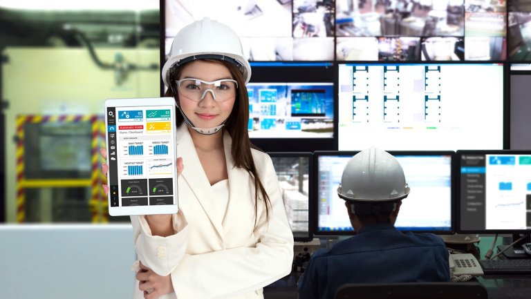 Female engineer wearing safety glasses and a white hard hat standing in front of a number of monitors and holding a tablet displaying a data dashboard