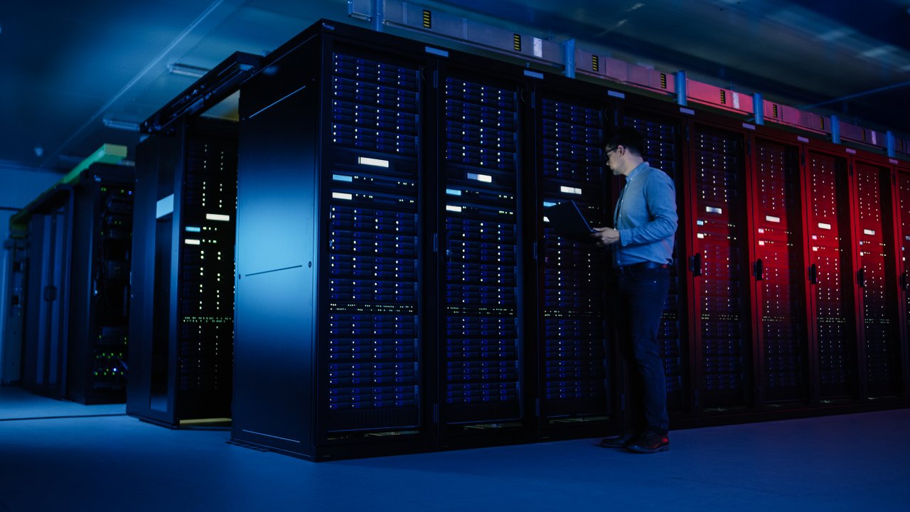 Um homem especialista em TI com camisa social caminha ao longo de uma fileira de racks de servidores operacionais usando um laptop para manutenção remota