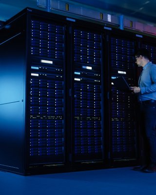 A male IT specialist in business shirt walks along a row of operating server racks using a laptop for remote maintenance