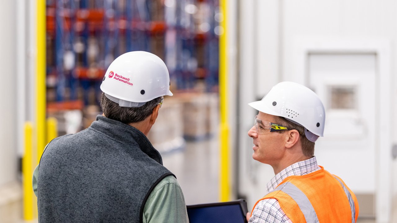 Consultor da Rockwell Automation usando um capacete observa um tablet junto a um cliente usando um colete laranja