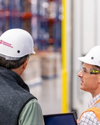 Rockwell Automation consultant in hard hat looks at tablet with male client in orange vest