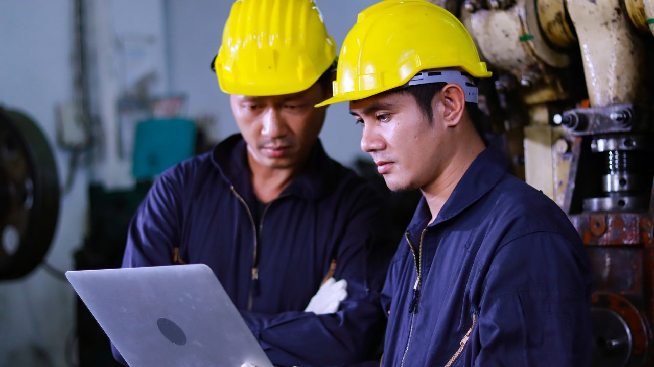 Two male maintenance engineers in safety clothing using laptops in a manufacturing plant.