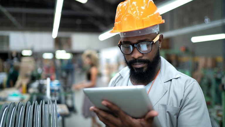 A man wearing glasses and a hard hat holding a tablet in a manufacturing plant