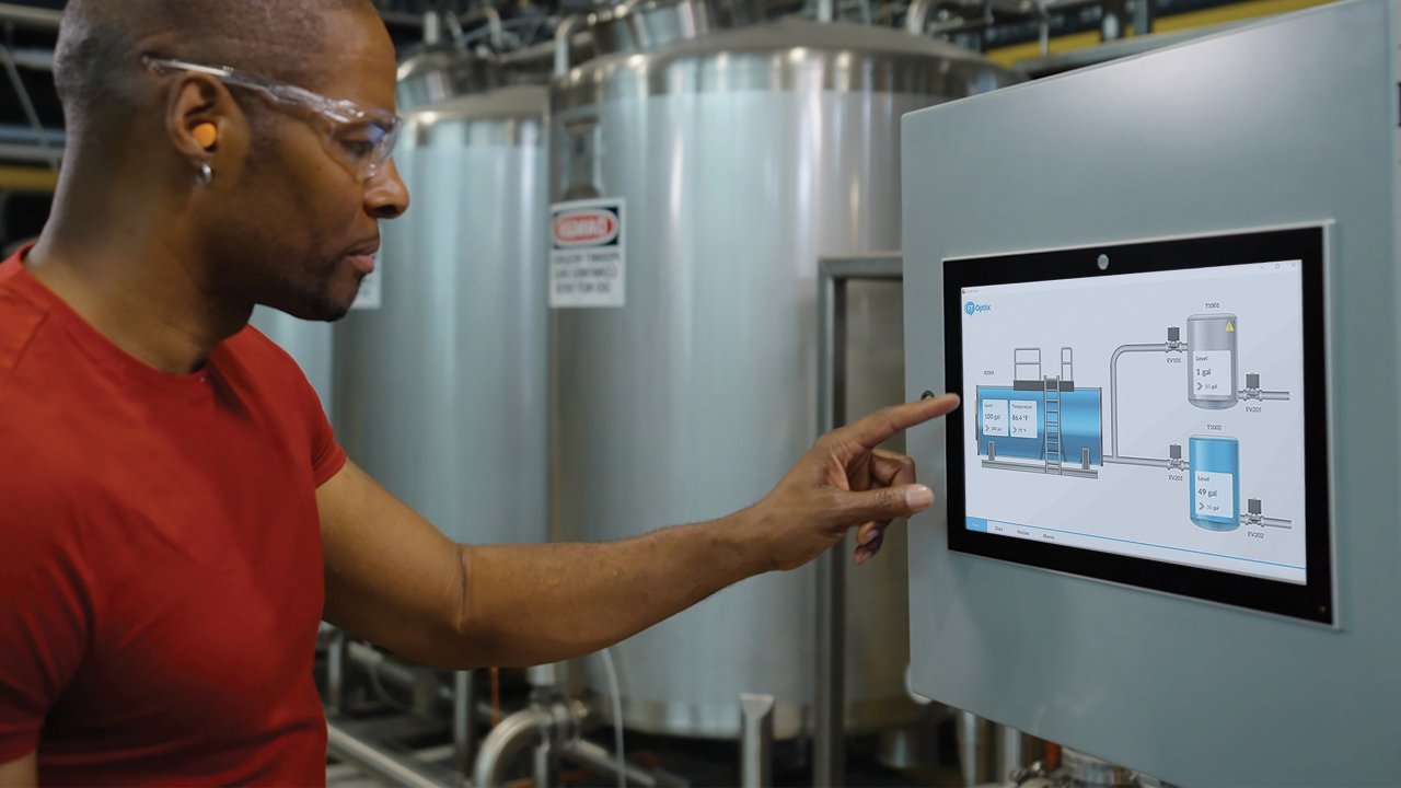 Homem apontando para uma tela touchscreen em um FactoryTalk OptixPanel, dentro de uma fábrica.