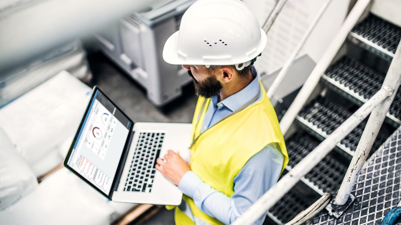 An industrial male engineer in a factory wearing a hardhat holding a laptop that displays the Fiix dashboard.