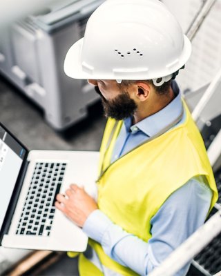 Um engenheiro industrial em uma fábrica, usando um capacete de segurança e segurando um laptop que exibe o painel Fiix.