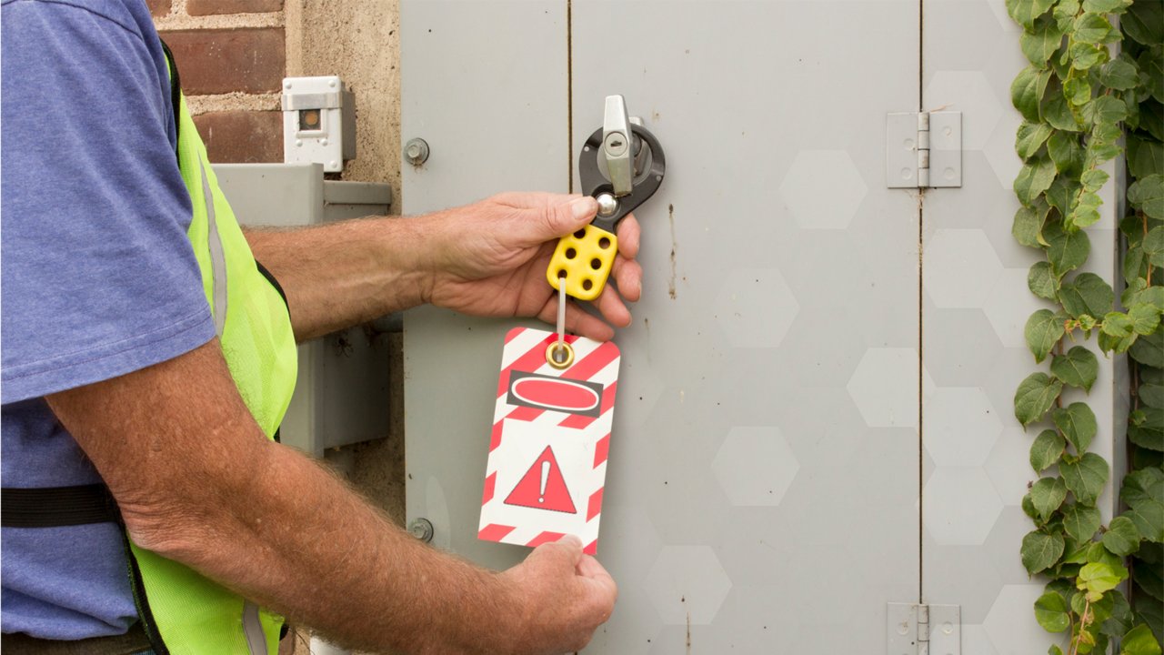 Facility worker in blue tshirt and yellow safety vest reviews lockout/tagout device recommendations on red and white diagnal striped tag with red triangle exclamation point in the middle below a black rectangle  covered by white oval and red oval hanging from cabinet door handle