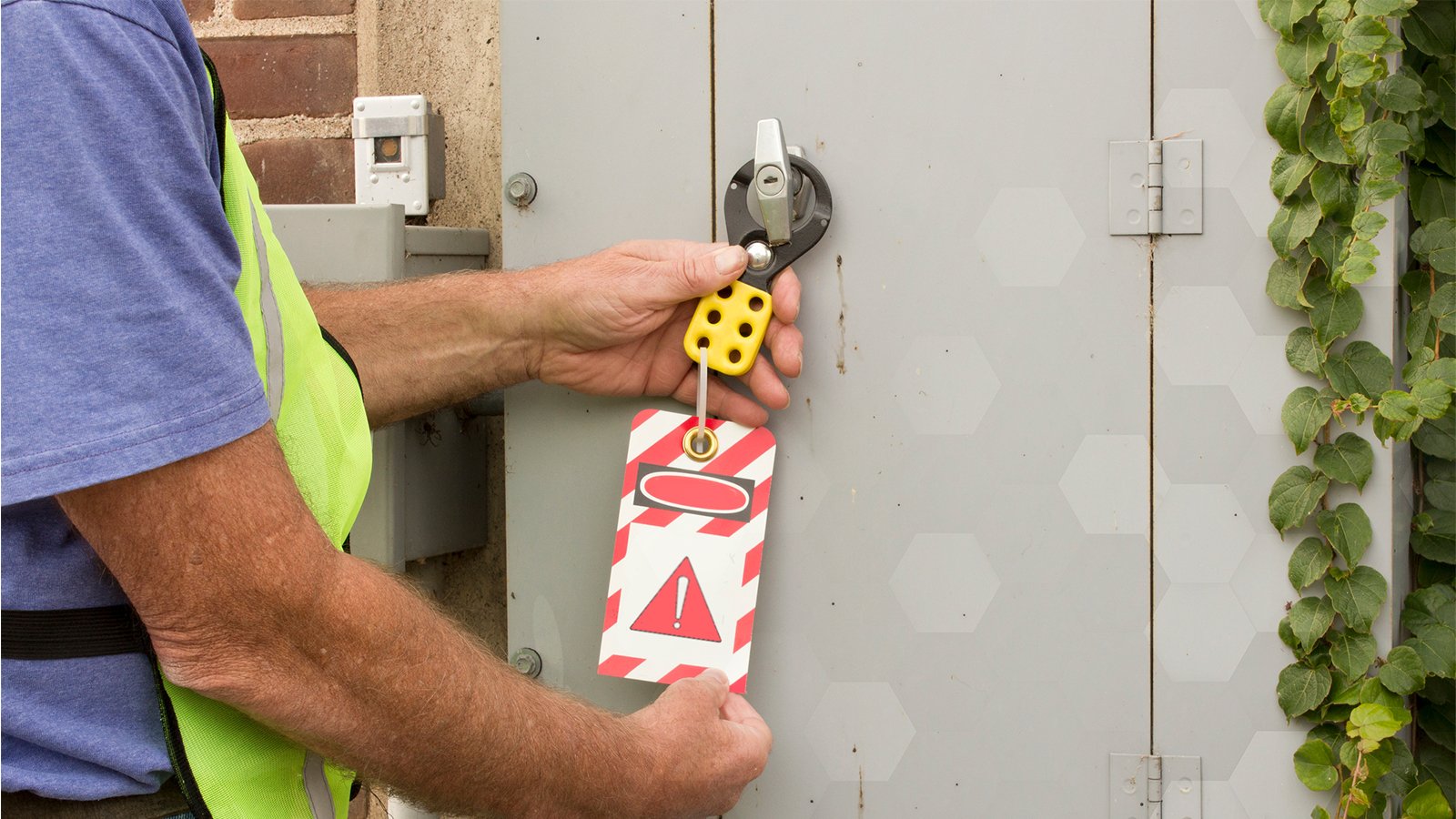 Employé d’usine portant une chemise bleue et un gilet jaune de sécurité examinant les recommandations en matière de dispositifs de verrouillage/signalisation inscrites sur une étiquette diagonale rayée rouge et blanche avec un point d’exclamation rouge dans un triangle noir, couverte d’un ovale blanc et d’un ovale rouge, suspendue sur la poignée du boîtier.