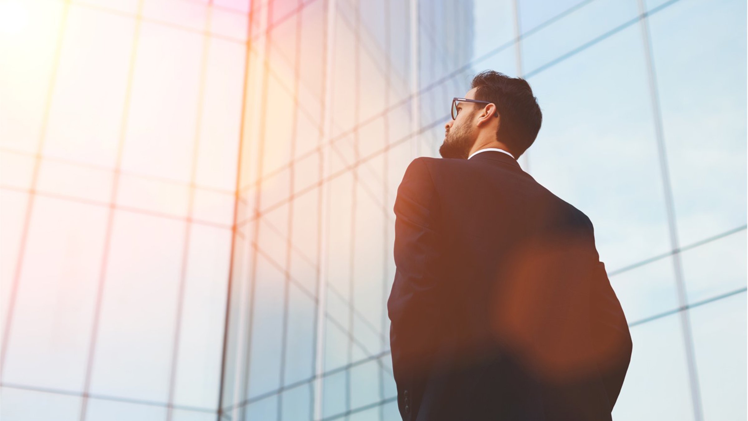 Man in dark suit with glasses looking at glass building with sun reflection