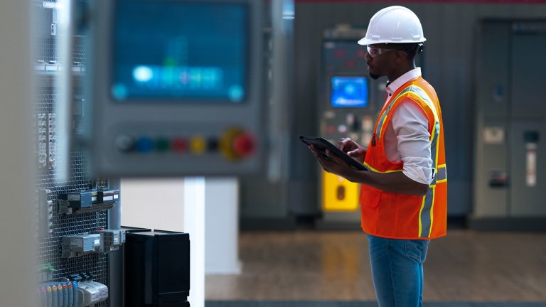 Man in protective safety glasses, vest and hard hat using tablet and looking at industrial machines