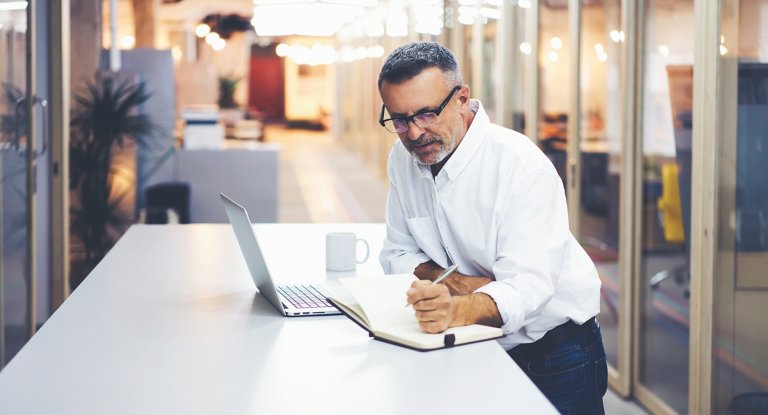 Homme portant des lunettes, debout à une table, prenant des notes dans un carnet, avec un ordinateur portable et une tasse de café sur la table