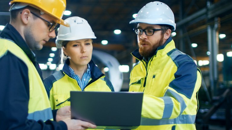 Três engenheiros usando capacetes e jaquetas de alta visibilidade olham para um laptop durante uma apreciação de risco de segurança de máquina.