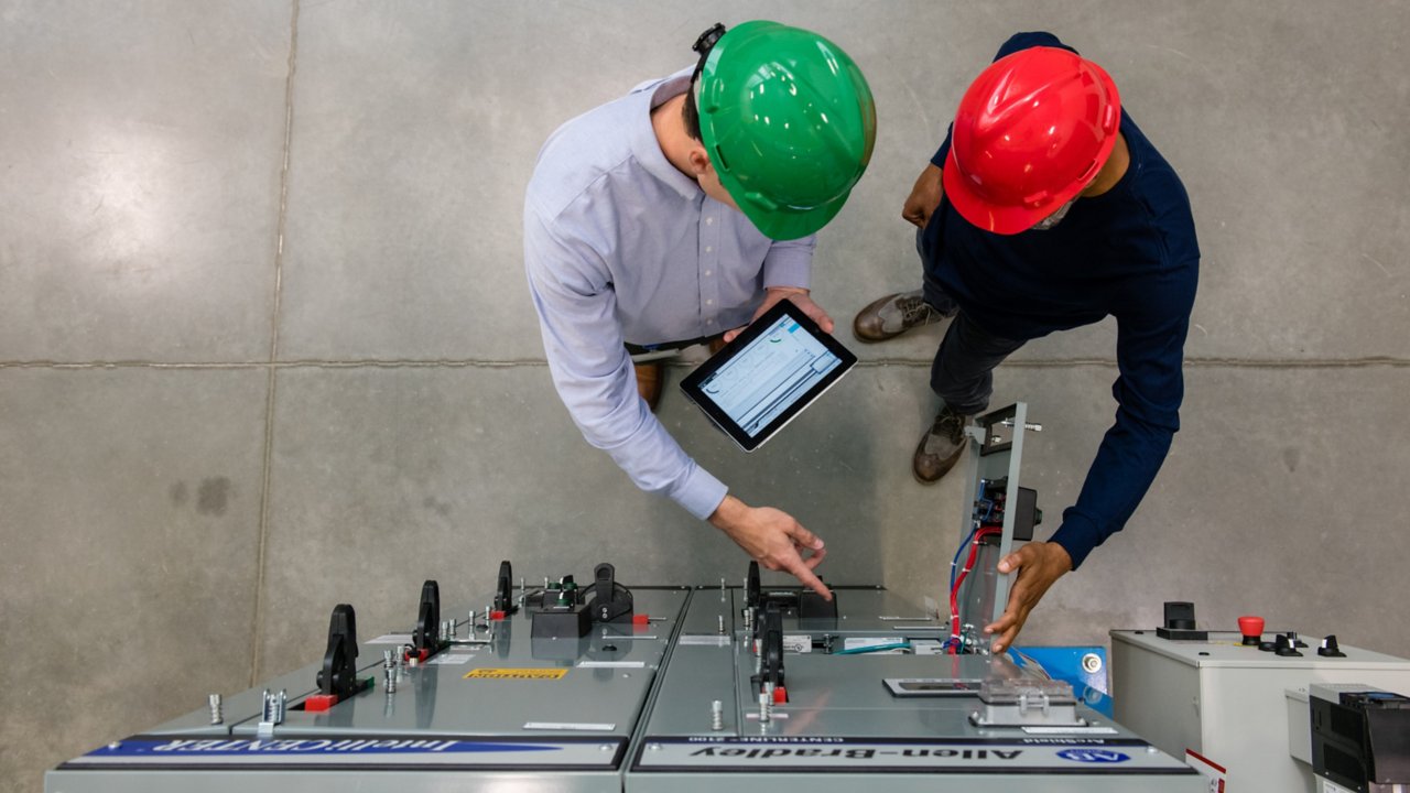 Two men in hard hats on the floor of a factory using a tablet while working with PlantPAX