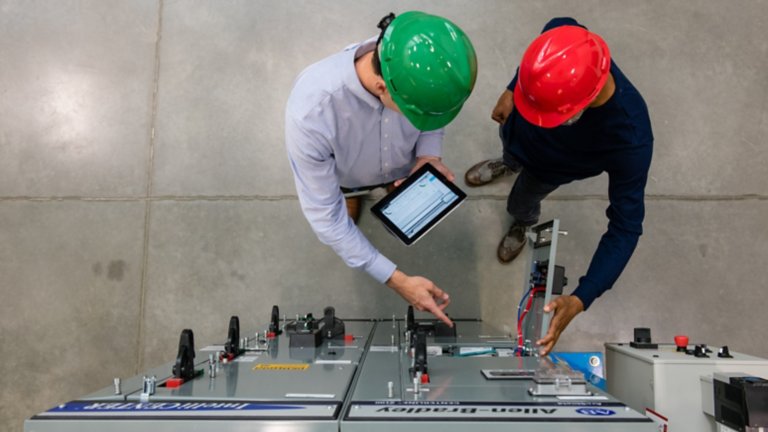 Two men in hard hats on the floor of a factory using a tablet while working with PlantPAX