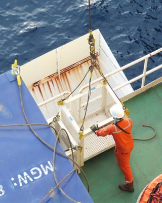 An oil and gas supply boat transfers cargo to a worker on deck ready with a hoisted basket. The worker is in an orange jumpsuit with a white safety helmet and gloves