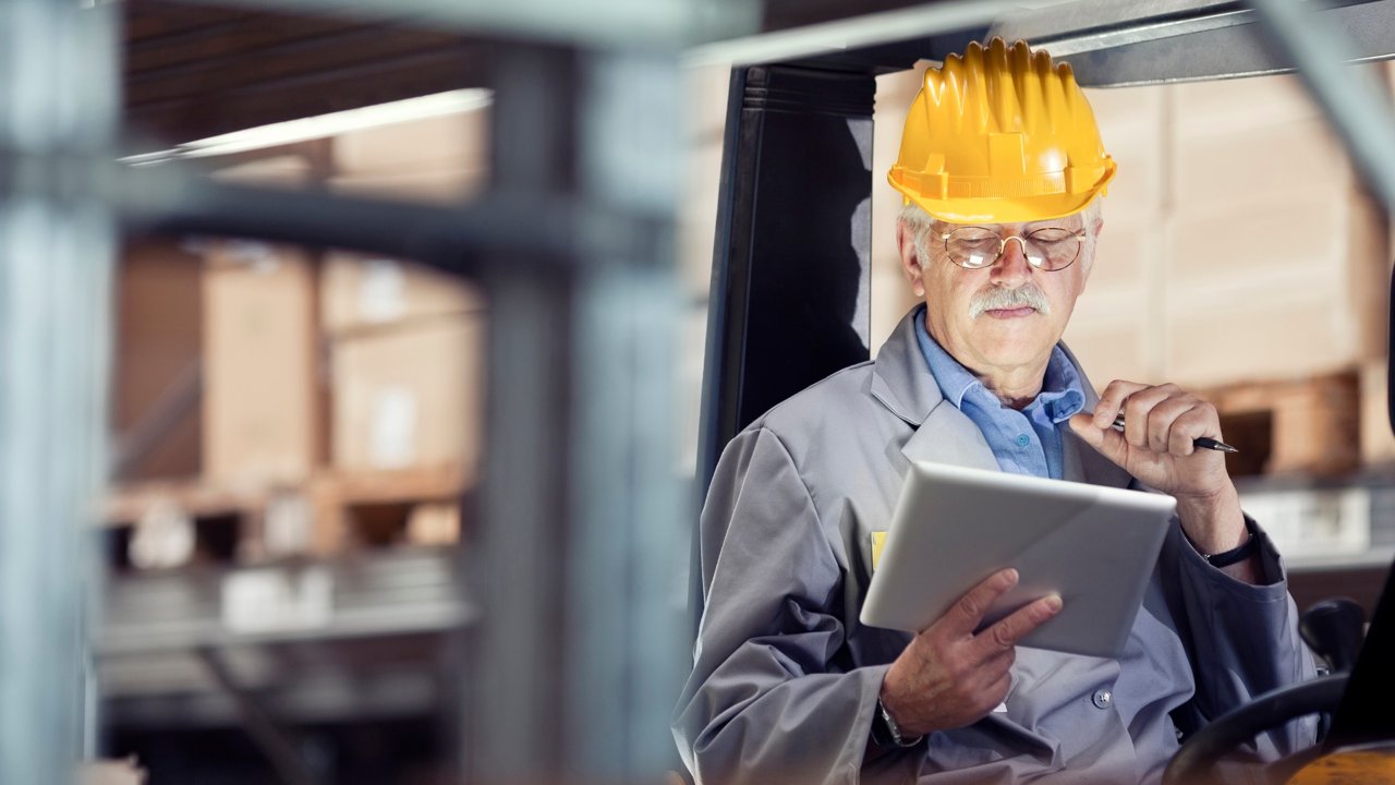 Un hombre con un casco y anteojos de seguridad con una tableta en la mano sentado en un elevador de horquilla en la planta