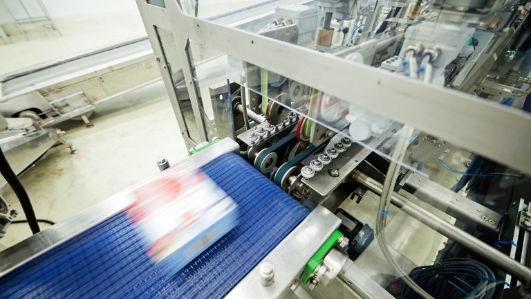 Una caja blanca con desenfoque de movimiento avanza a toda velocidad por una cinta transportadora azul hacia una máquina de producción.