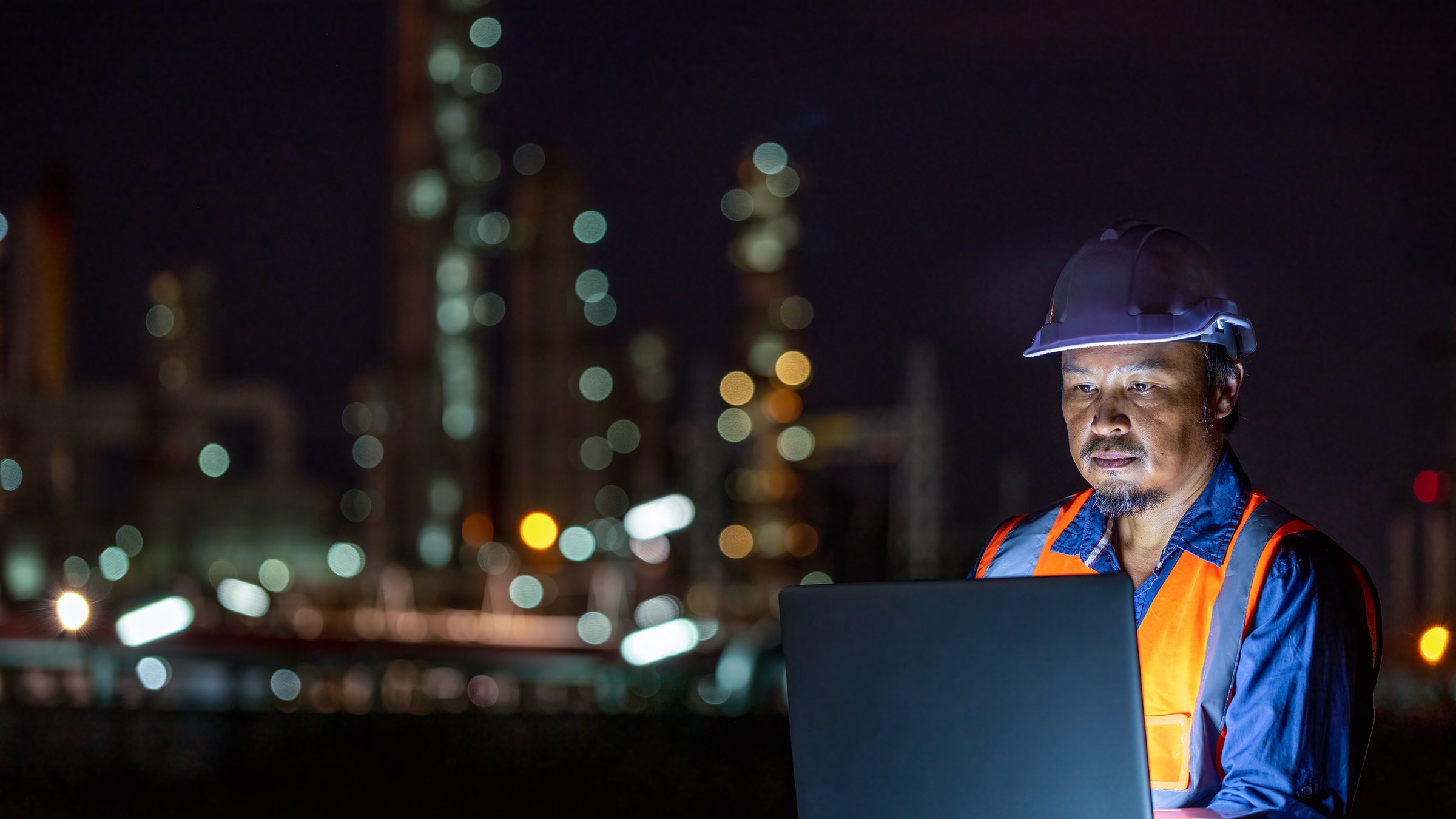 Un trabajador usa una computadora portátil con una planta petroquímica en la noche en el fondo.