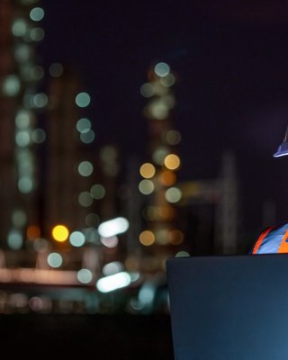 Un trabajador usa una computadora portátil con una planta petroquímica en la noche en el fondo.