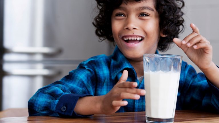 微笑着的男孩坐在一张桌子旁，端着一杯牛奶