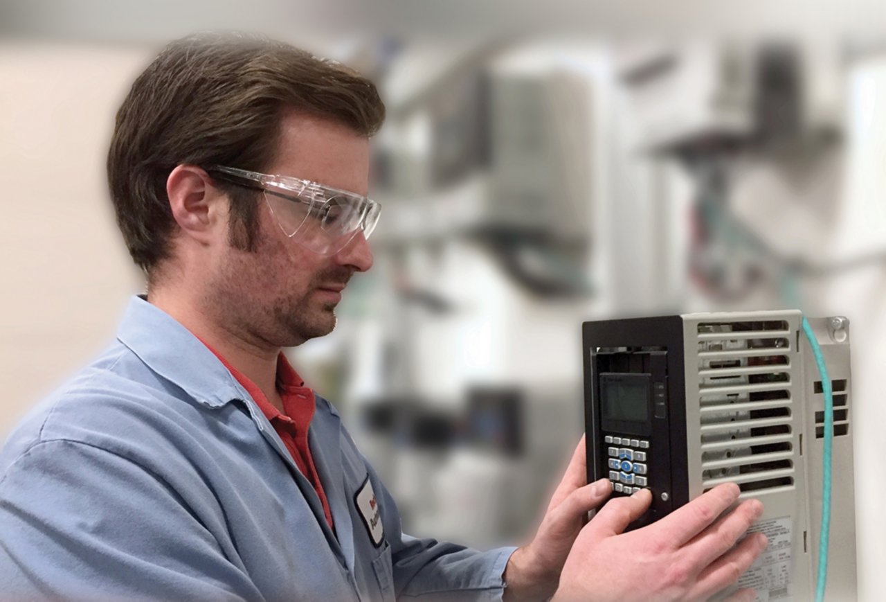 Un hombre con anteojos de seguridad y bata de laboratorio azul instalando un variador de frecuencia PowerFlex, El variador de frecuencia está a la derecha y es gris, el abd rectangular tiene botones en una cara negra.