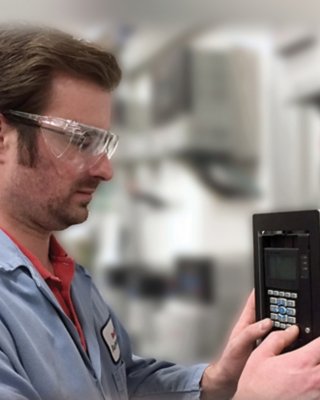 Male with safety glasses and blue lab coat installing a PowerFlex VFD, The VFD is on the right and is gray, rectangular abd has buttons on a black face.