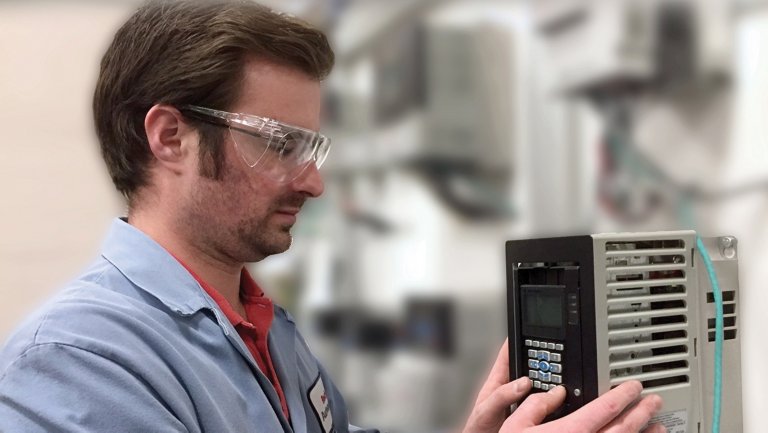 Male with safety glasses and blue lab coat installing a PowerFlex VFD, The VFD is on the right and is gray, rectangular abd has buttons on a black face.