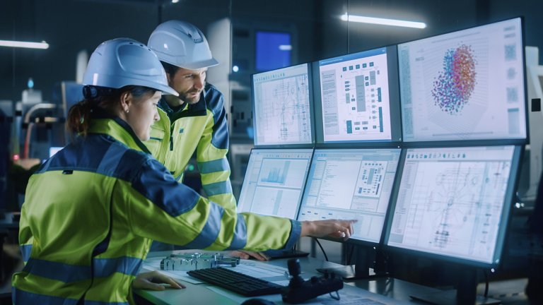 Two manufacturing employees wearing hard hats discuss operations while looking at multiple computer screens displaying production data.