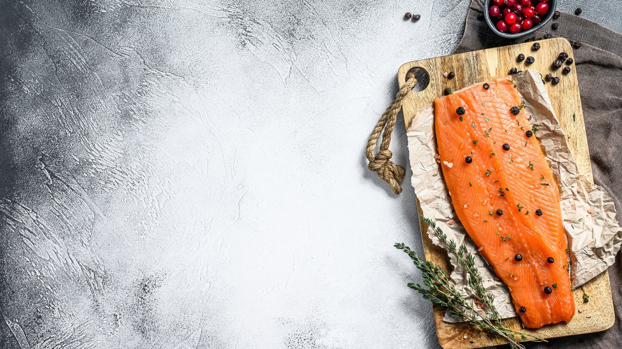 Ein Stück Lachs auf einem Schneidebrett mit Kräutern auf der linken Hälfte und eine Tasse Beeren darüber.