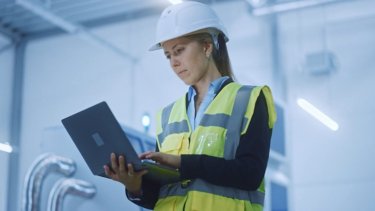 Female worker in hard hat and safety vest using a laptop on the job site.